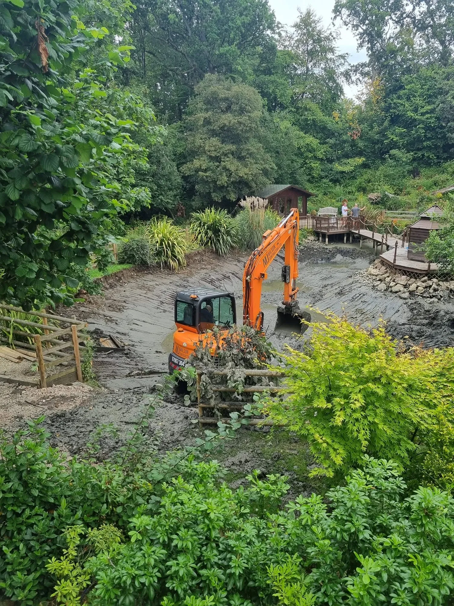 Excavator working in a drained garden pond surrounded by trees