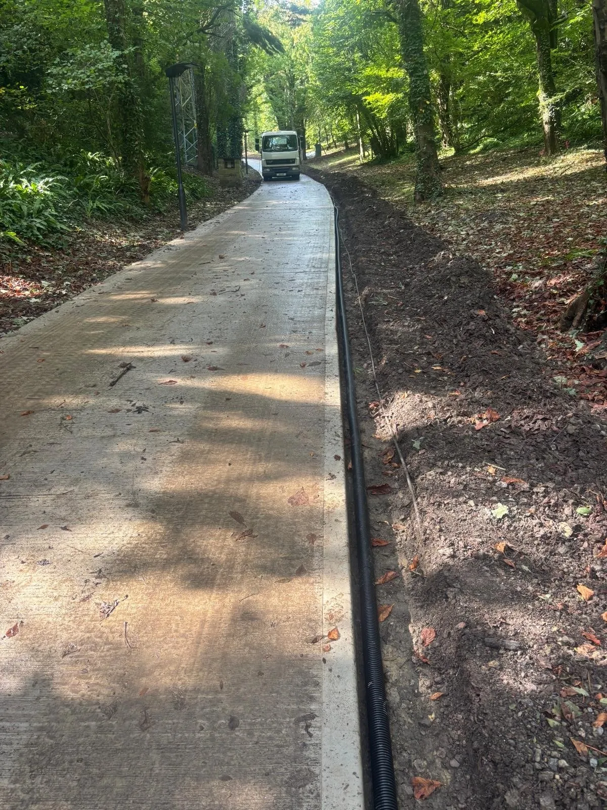 Finished concrete access road curving through woodland with ducting at the verge