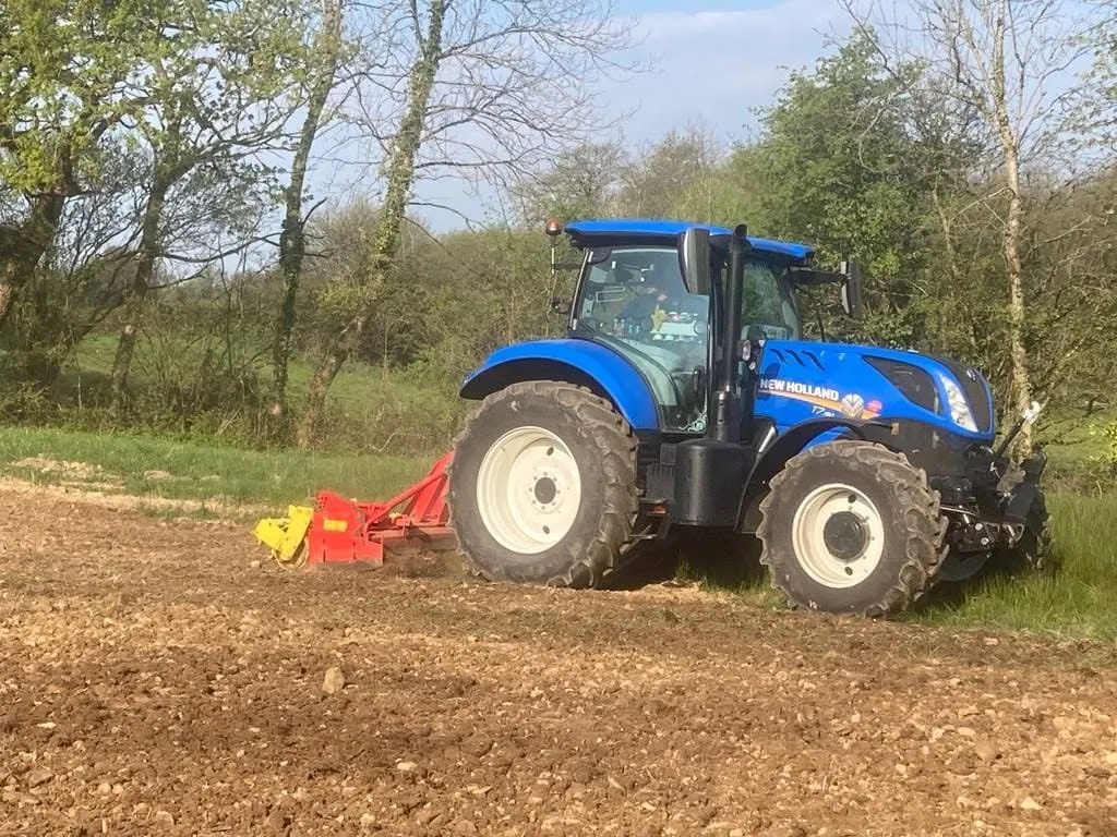 Tractor with a power harrow working a field beside woodland