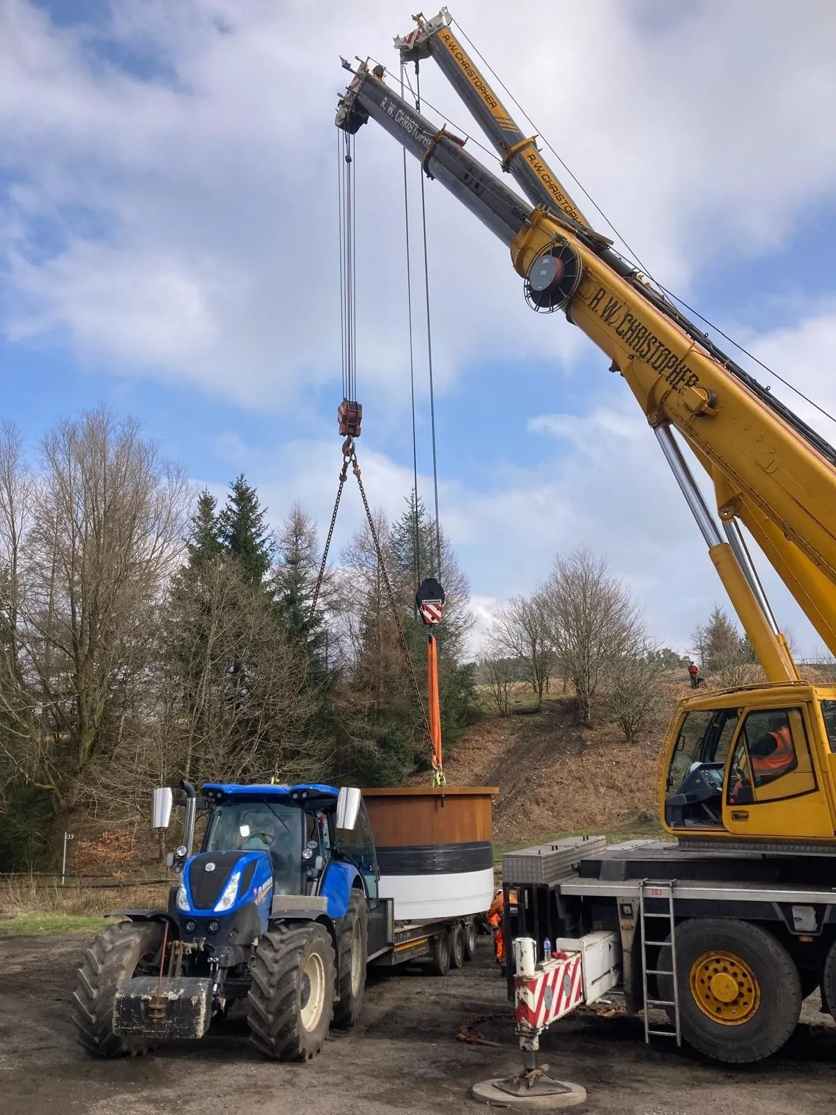 Crane lifting a tank from a low loader beside a New Holland tractor