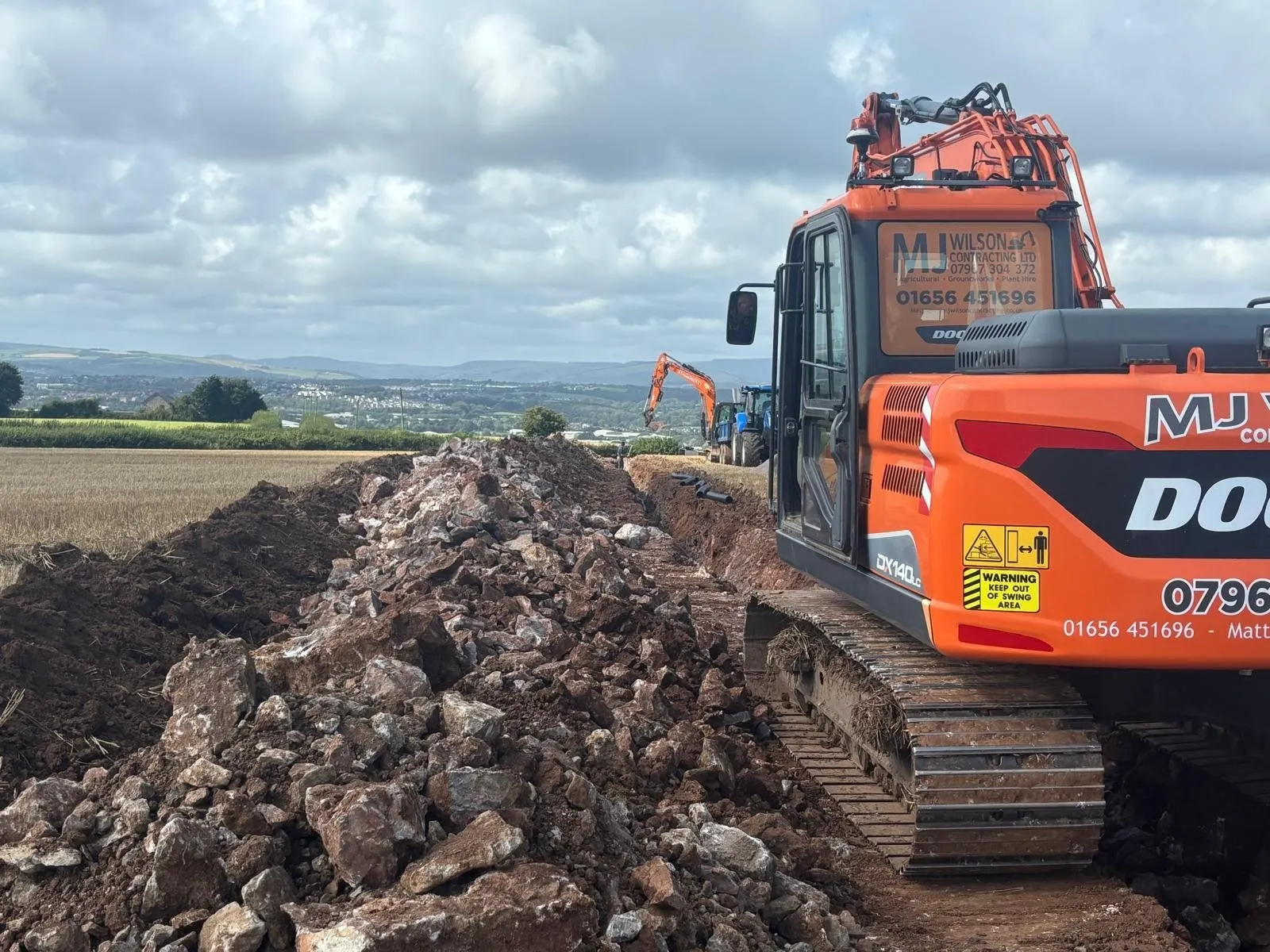 DX140LC excavator beside a rock-filled trench with hills behind