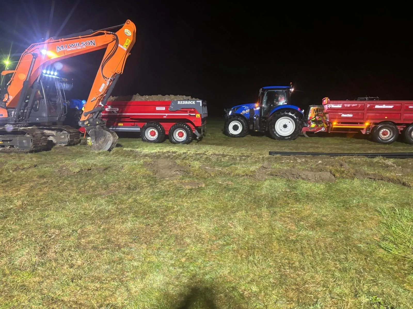 Excavator loading a dumper trailer at night under work lights, with a tractor and trailer waiting