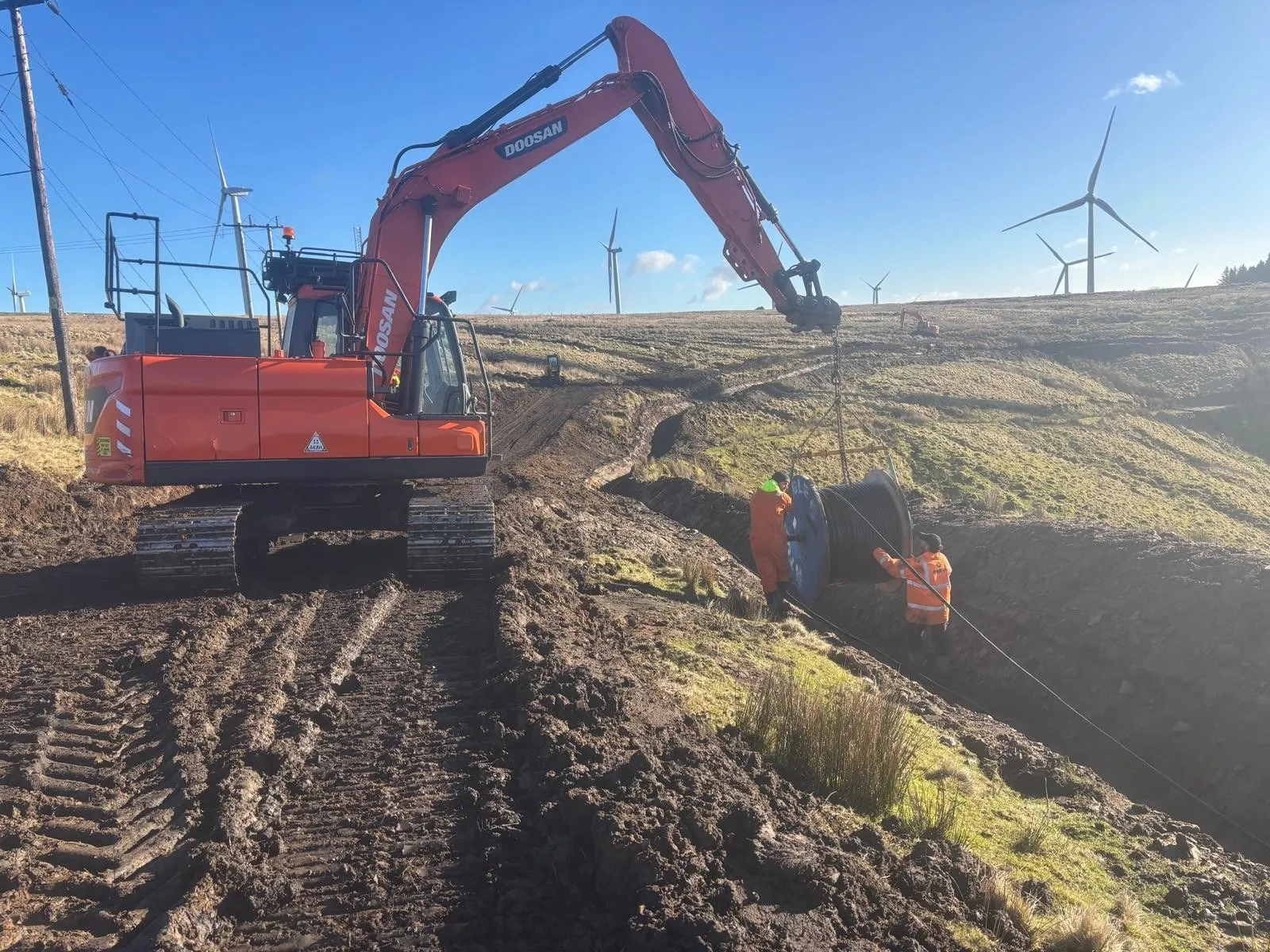 Excavator holding a cable drum while two operatives pull cable into a trench on a wind farm