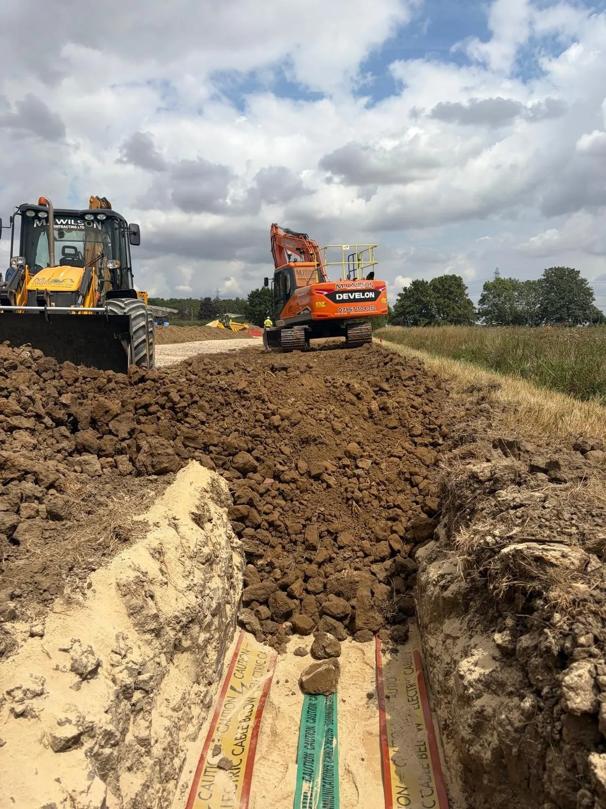 JCB backhoe and Develon excavator backfilling a trench over cable warning tape